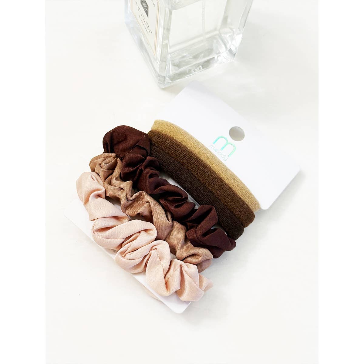 The Love and Repeat Assorted Hair Tie & Scrunchie Set, featuring four scrunchies in shades of brown and beige, is displayed on a white card next to a clear glass bottle on a white surface.