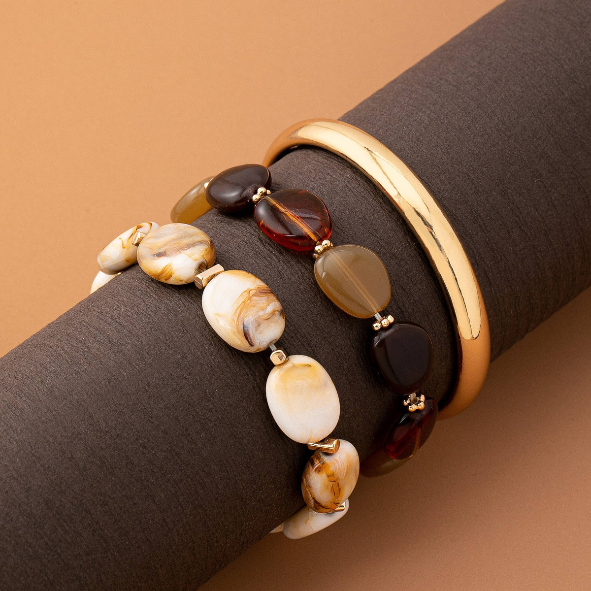 The Boho Gold Bracelet Set by JOSSLYN by wall to wall is showcased on a dark brown cylindrical stand, featuring a smooth gold bangle, a polished bracelet with multicolored stones, and another adorned with marbled cream and tan beads, all set against a light brown background.