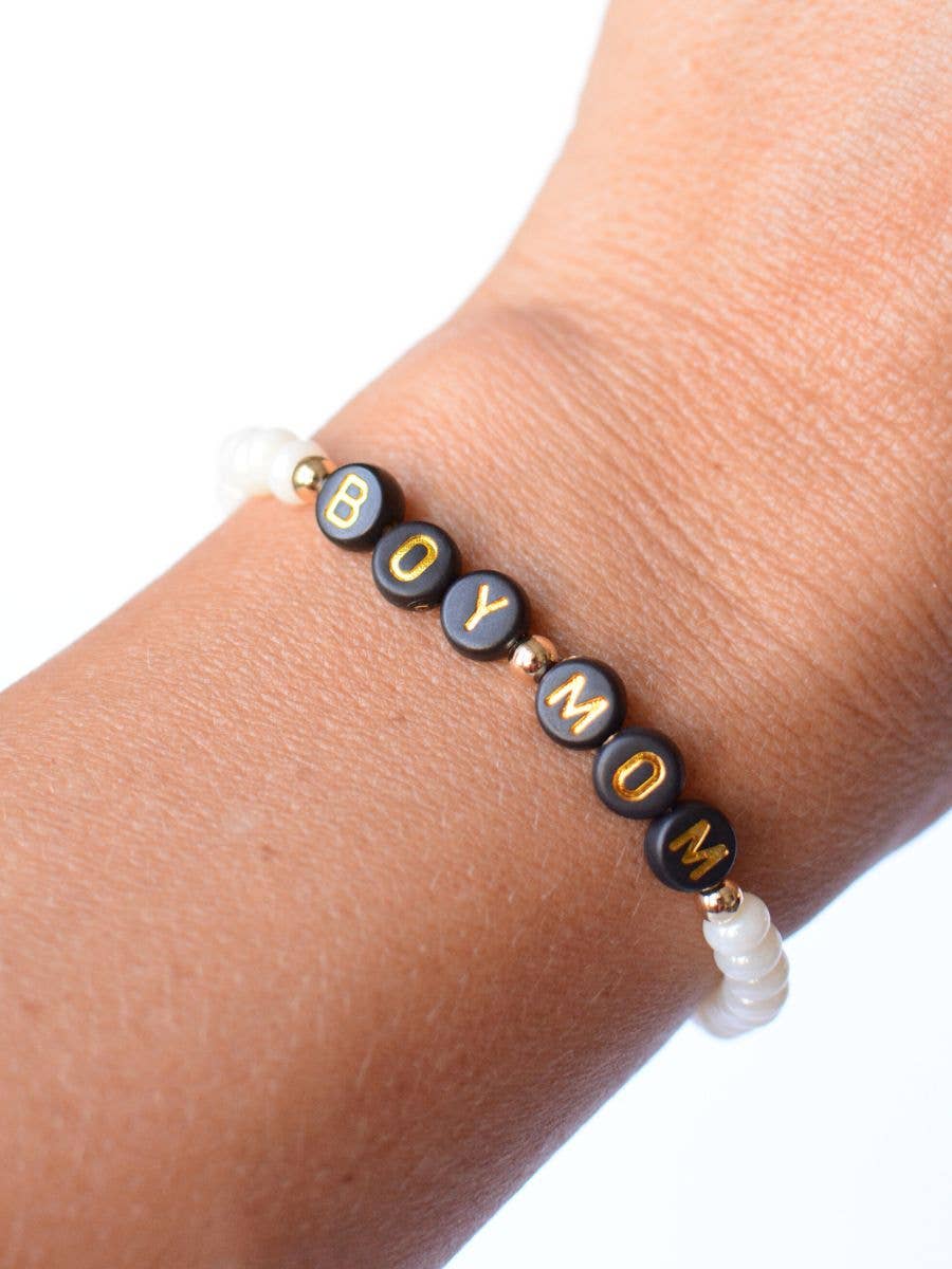 A close-up of a person’s wrist wearing the Boy Mom Pearl Bracelet by Two and Crew - Faith Based Jewelry & Gifts, featuring black round beads that spell “BOY MOM” in gold letters, separated by small gold and white beads.