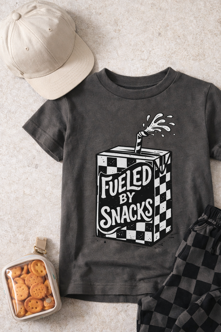 A Raising Brave Fueled By Snacks Toddler Tee in gray, featuring a juice box graphic and text, is paired with a beige cap, black checkered shorts, and a small container of cheese crackers.