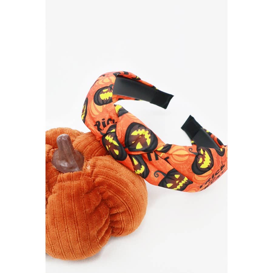 Halloween Theme Headband featuring an orange pattern of jack-o-lanterns and the words trick or treat, resting next to a plush orange pumpkin.