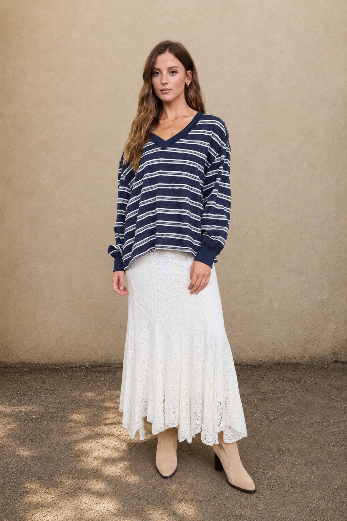 A person wears a Blu Pepper Blue Stripe V-Neck Long Sleeve navy blue shirt with horizontal white stripes, a long white lace skirt, and tan ankle boots.