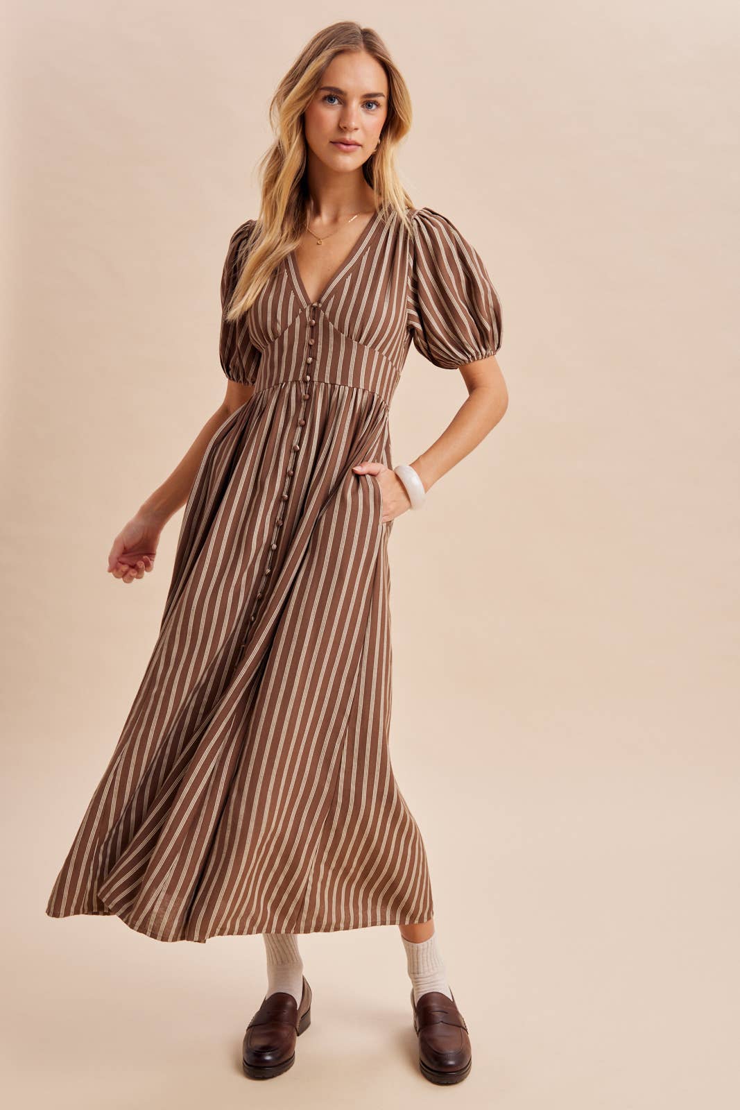 A woman wears an In Loom Stripe Button Down Maxi Dress featuring brown and white stripes, puff sleeves, a V-neck, and a button-front closure.