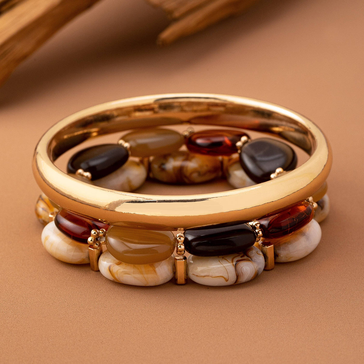 The Boho Gold Bracelet Set by JOSSLYN by wall to wall features a stylish gold bangle adorned with oval and round stones in brown, black, red, and cream tones, beautifully displayed on a neutral tan background.