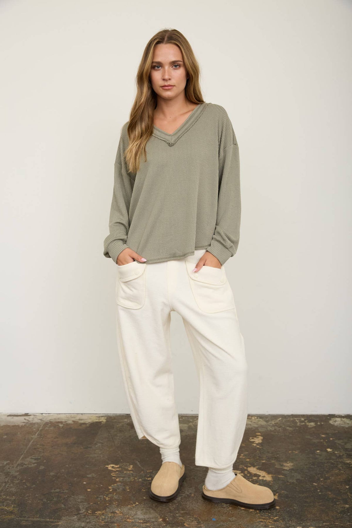 A woman with long hair wears the Blu Pepper Olive V-Neck Long Sleeve Top and white pants with large front pockets. She stands against a plain wall with her hands in her pockets, wearing tan slippers on a textured floor.