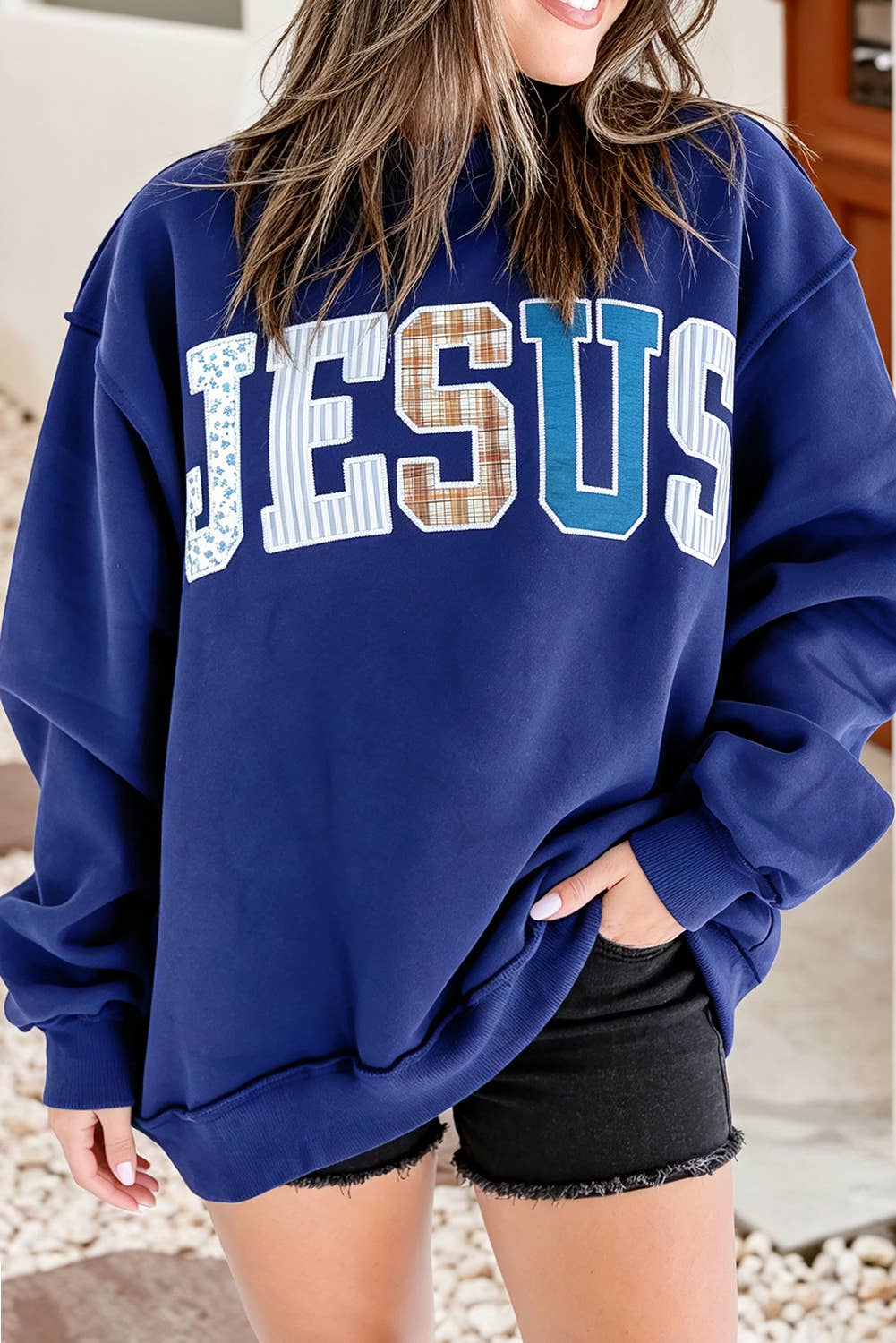 A navy blue Little Daisy Closet Jesus Mock Neck Sweatshirt with the word JESUS spelled out in large, multi-patterned fabric letters across the chest.