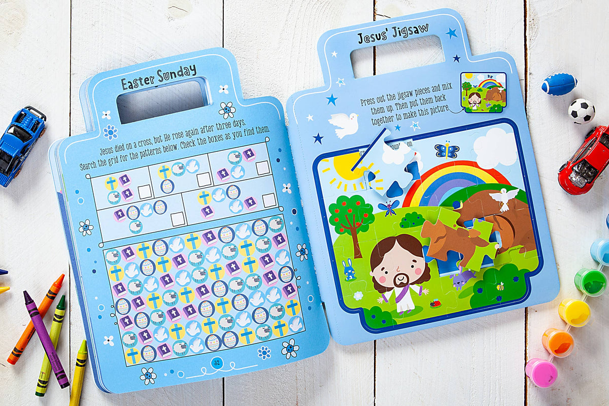 The My Bible Backpack Activity Book by BroadStreet Publishing Group, LLC is open on a wooden table, surrounded by crayons and toy cars. The pages feature a word search and a Jesus-themed jigsaw puzzle, decorated with colorful illustrations of Jesus, animals, and a rainbow.