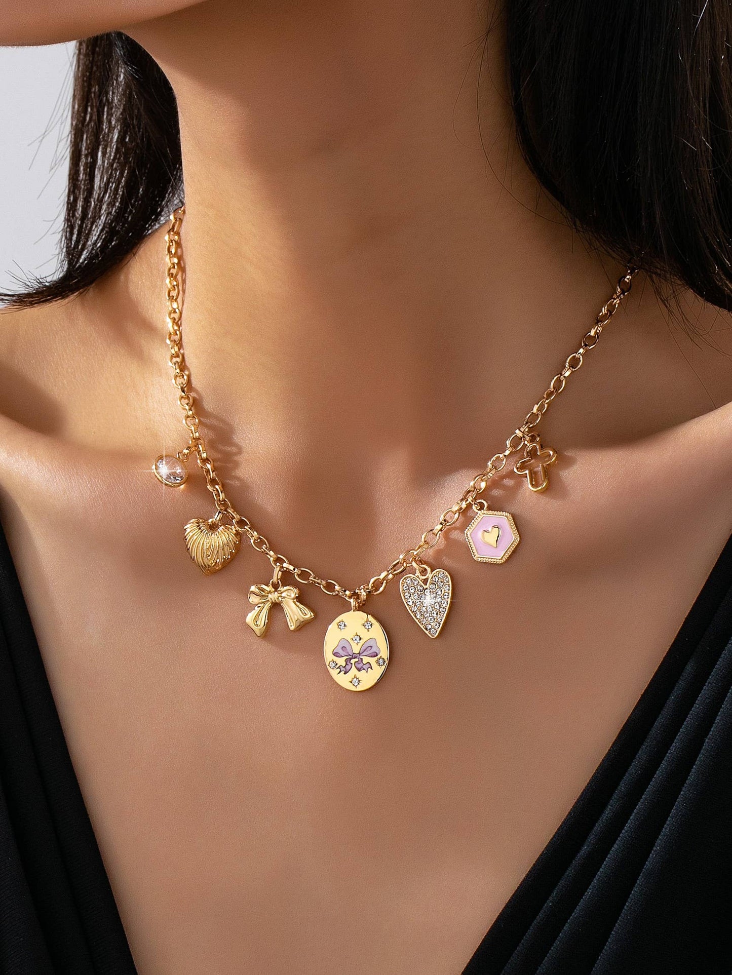 A close-up of a person wearing the JOSSLYN by wall to wall Pink Multi-Charm Necklace, featuring assorted gold pendants including a heart, star, seashell, angel, and round charms, styled over a black v-neck top.