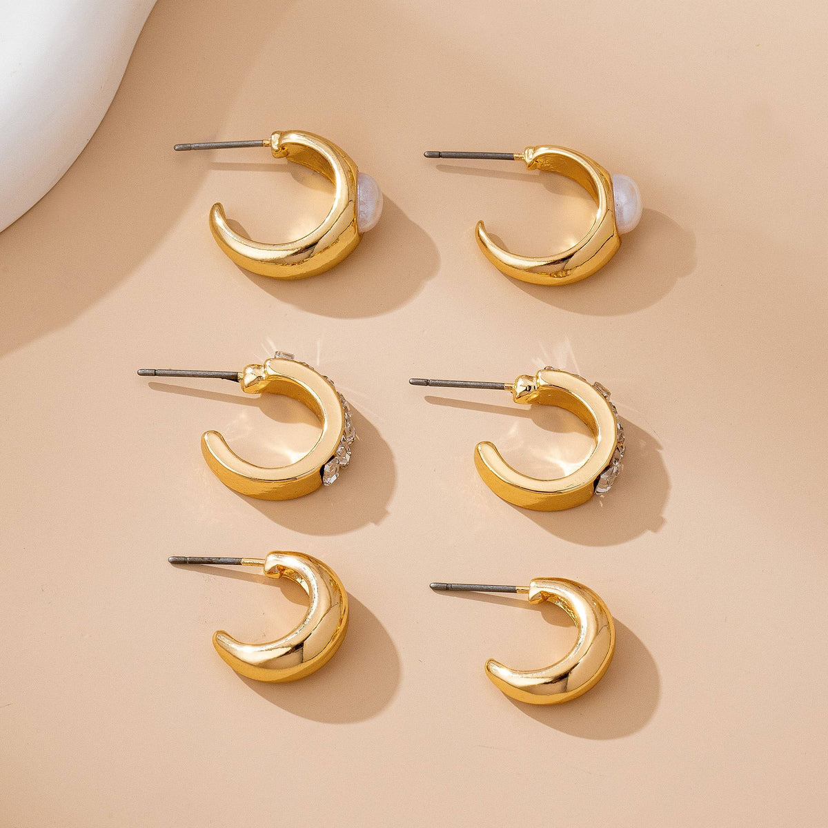 Six gold crescent moon-shaped earrings from the JOSSLYN by wall to wall Pearl & Baguette Hoop Trio Earring Set, featuring accents like pearls and small crystals, are arranged in two rows on a beige surface.