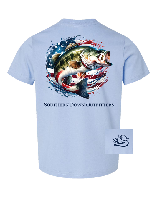 The American Bass Toddler Tee by Southern Down Outfitters is a light blue shirt with a bold bass fish over an American flag graphic on the back, brand text below, and a small hook logo in the lower corner.